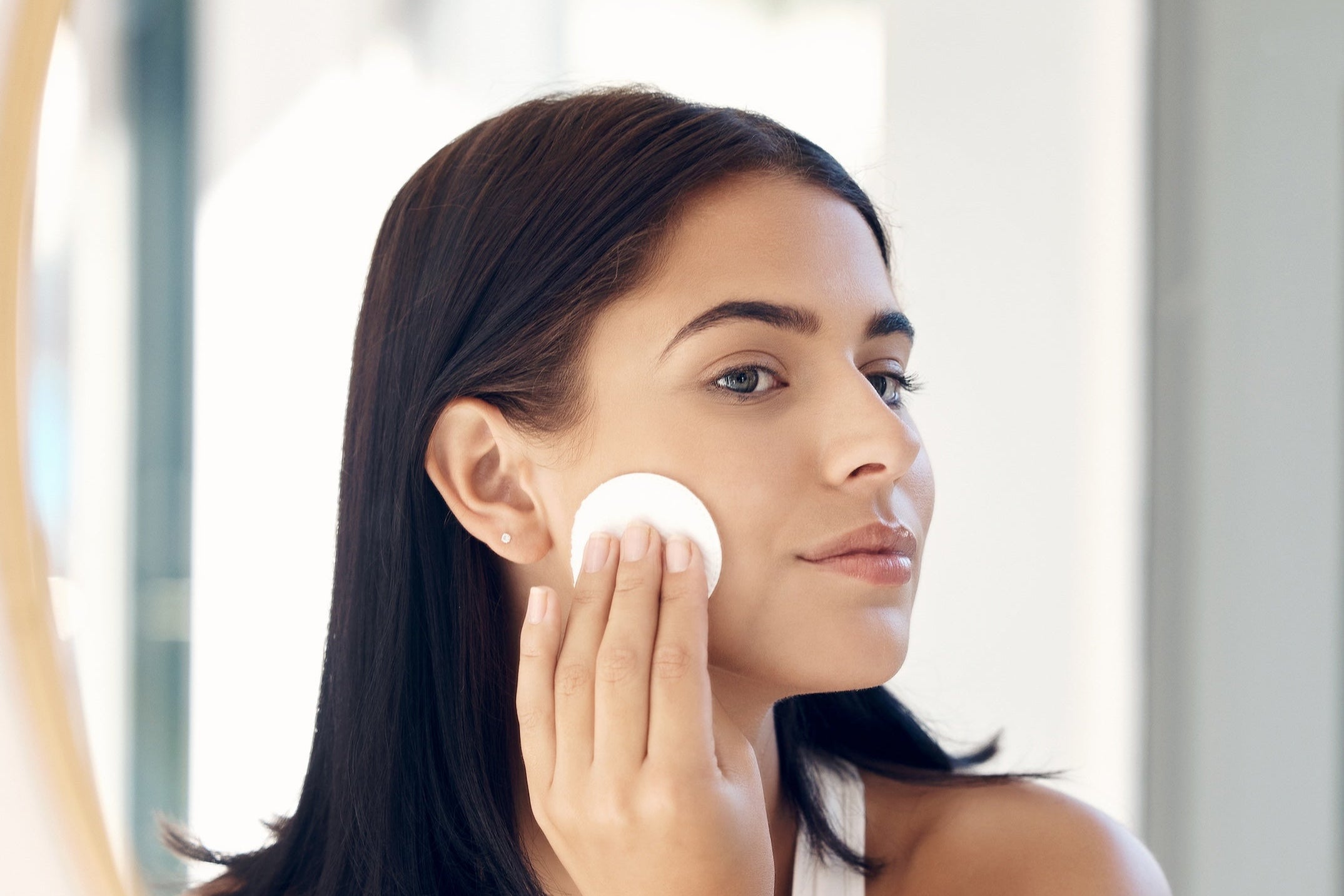 Mirror reflection of a woman wiping her face with cotton pad for acne control.
